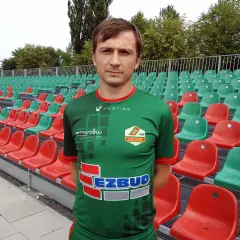 Kamil Szymczak w zielonej koszulce sportowej stoi przed rzędem czerwonych i zielonych krzesełek na stadionie.