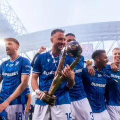 Piłkarze w niebieskich strojach świętują z pucharem i piłką na stadionie.