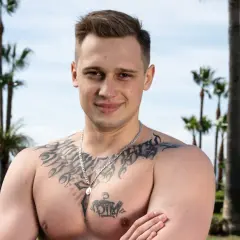 Daniel Majewski, młody mężczyzna z tatuażami na klatce piersiowej, pozuje na tle palm i błękitnego nieba.