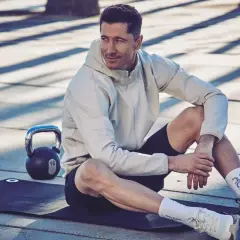 Robert Lewandowski w sportowym stroju, siedzi na macie treningowej obok odważnika. Ciekawe informacje o jego karierze i treningach.