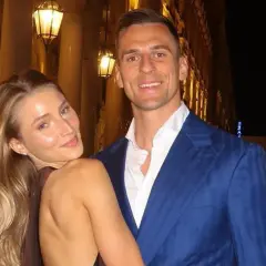Uśmiechnięta dziewczyna z blond włosami opiera się o mężczyznę w niebieskiej marynarce. Spacerują wieczorem ulicą z latarniami.