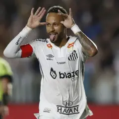 Neymar w białej koszulce Santos z czerwonymi opaskami na ramionach, pokazuje język i robi śmieszne miny.