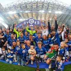 Byli piłkarze Lecha Poznań świętują z pucharem i dziećmi na murawie stadionu.