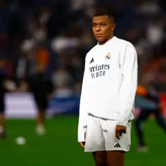 Mbappé, młody mbp piłkarz, w białej koszulce i spodenkach Realu Madryt, stoi na murawie stadionu.