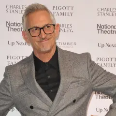 Gary Lineker w szarej marynarce i okularach, uśmiecha się na tle ścianki z logo National Theatre.