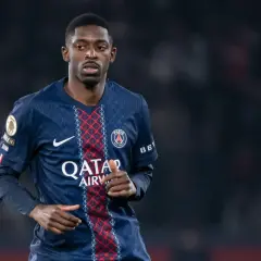 Piłkarz PSG, gdzie gra Dembele, w ciemnoniebieskiej koszulce z czerwonymi i białymi detalami, na tle rozmytego stadionu.