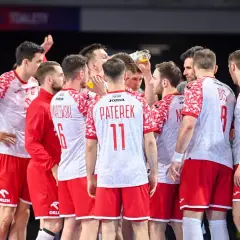 Drużyna piłkarzy ręcznych Polski w czerwono-białych strojach w skupieniu słucha trenera.