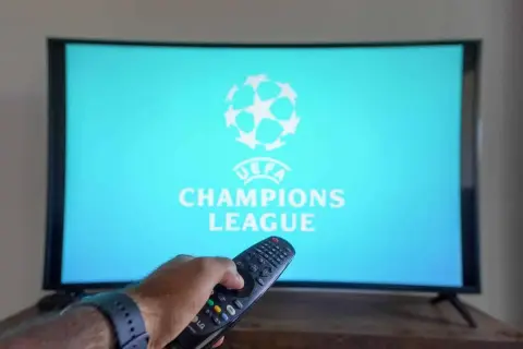 Ręka z pilotem szuka, gdzie transmisja Ligi Mistrzów. Na ekranie logo UEFA Champions League.