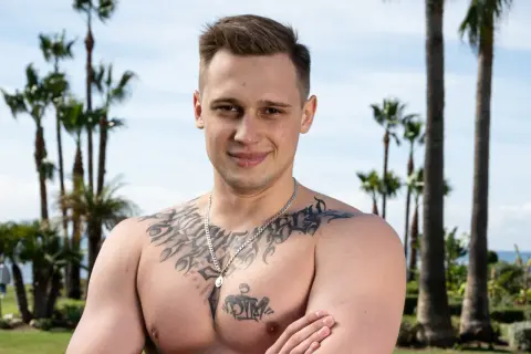 Daniel Majewski, młody mężczyzna z tatuażami na klatce piersiowej, pozuje na tle palm i błękitnego nieba.
