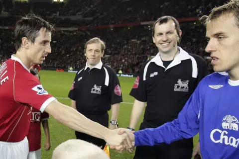 Piłkarz w czerwonej koszulce, Gary Neville, ściska dłoń rywalowi. Sędziowie obserwują.