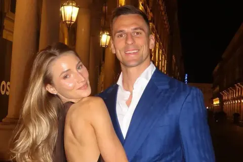 Uśmiechnięta dziewczyna z blond włosami opiera się o mężczyznę w niebieskiej marynarce. Spacerują wieczorem ulicą z latarniami.