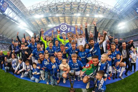 Byli piłkarze Lecha Poznań świętują z pucharem i dziećmi na murawie stadionu.
