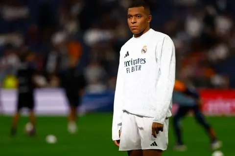 Mbappé, młody mbp piłkarz, w białej koszulce i spodenkach Realu Madryt, stoi na murawie stadionu.