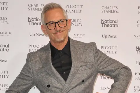 Gary Lineker w szarej marynarce i okularach, uśmiecha się na tle ścianki z logo National Theatre.