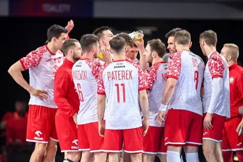 Drużyna piłkarzy ręcznych Polski w czerwono-białych strojach w skupieniu słucha trenera.
