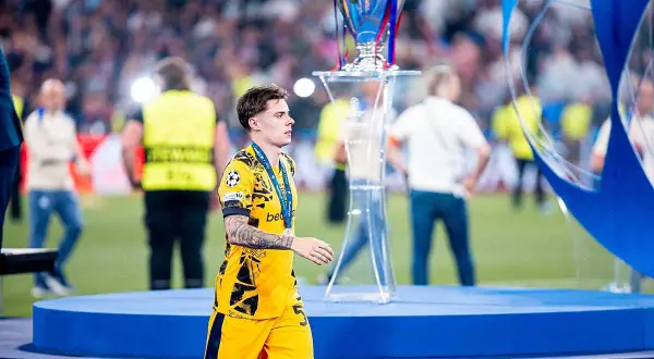 Piłkarz w żółtej koszulce, Nicola Zalewski, z medalem na szyi, idzie obok pucharu Ligi Mistrzów. Jego wiek to 22 lata.