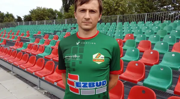 Kamil Szymczak w zielonej koszulce sportowej stoi przed rzędem czerwonych i zielonych krzesełek na stadionie.
