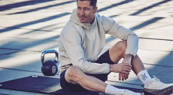 Robert Lewandowski w sportowym stroju, siedzi na macie treningowej obok odważnika. Ciekawe informacje o jego karierze i treningach.