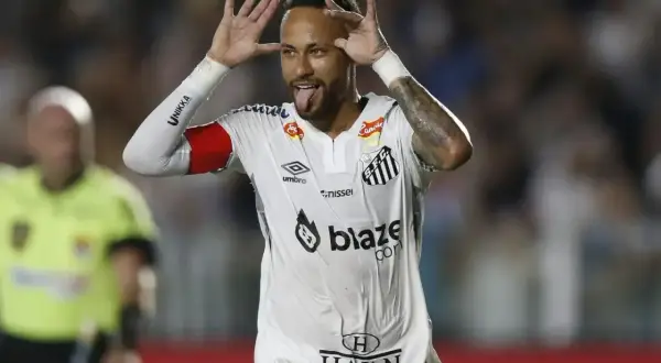 Neymar w białej koszulce Santos z czerwonymi opaskami na ramionach, pokazuje język i robi śmieszne miny.