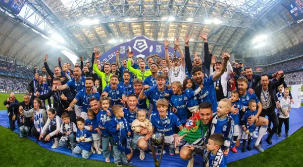Byli piłkarze Lecha Poznań świętują z pucharem i dziećmi na murawie stadionu.