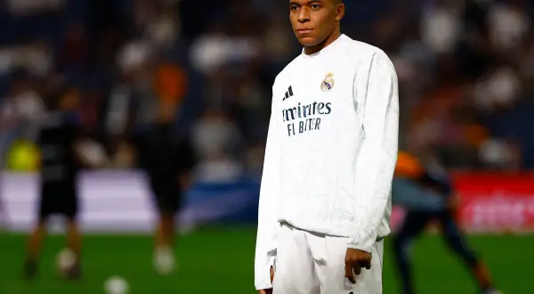 Mbappé, młody mbp piłkarz, w białej koszulce i spodenkach Realu Madryt, stoi na murawie stadionu.