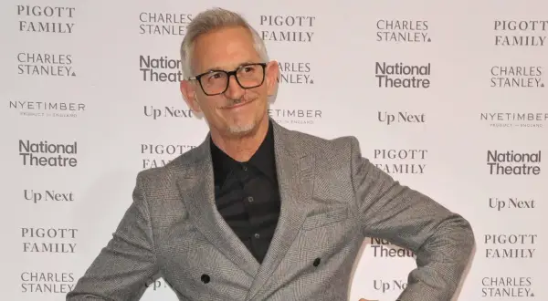 Gary Lineker w szarej marynarce i okularach, uśmiecha się na tle ścianki z logo National Theatre.