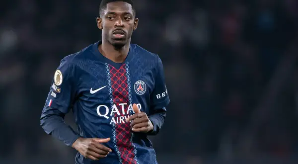 Piłkarz PSG, gdzie gra Dembele, w ciemnoniebieskiej koszulce z czerwonymi i białymi detalami, na tle rozmytego stadionu.