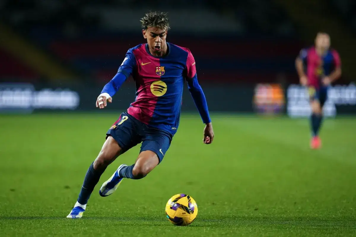 Młody piłkarz FC Barcelony w bordowo-niebieskiej koszulce z numerem 7 drybluje z piłką na zielonej murawie.