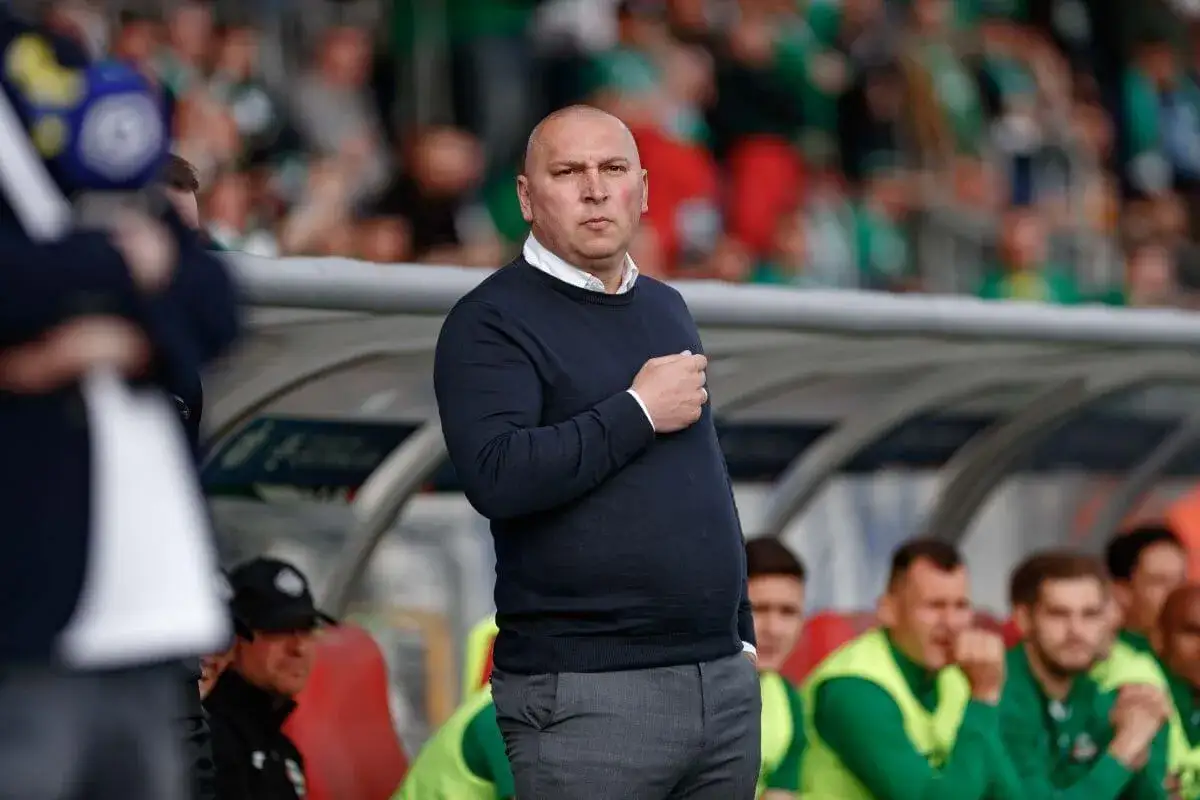 Mariusz Lewandowski, trener w granatowym swetrze, stoi na tle trybun.