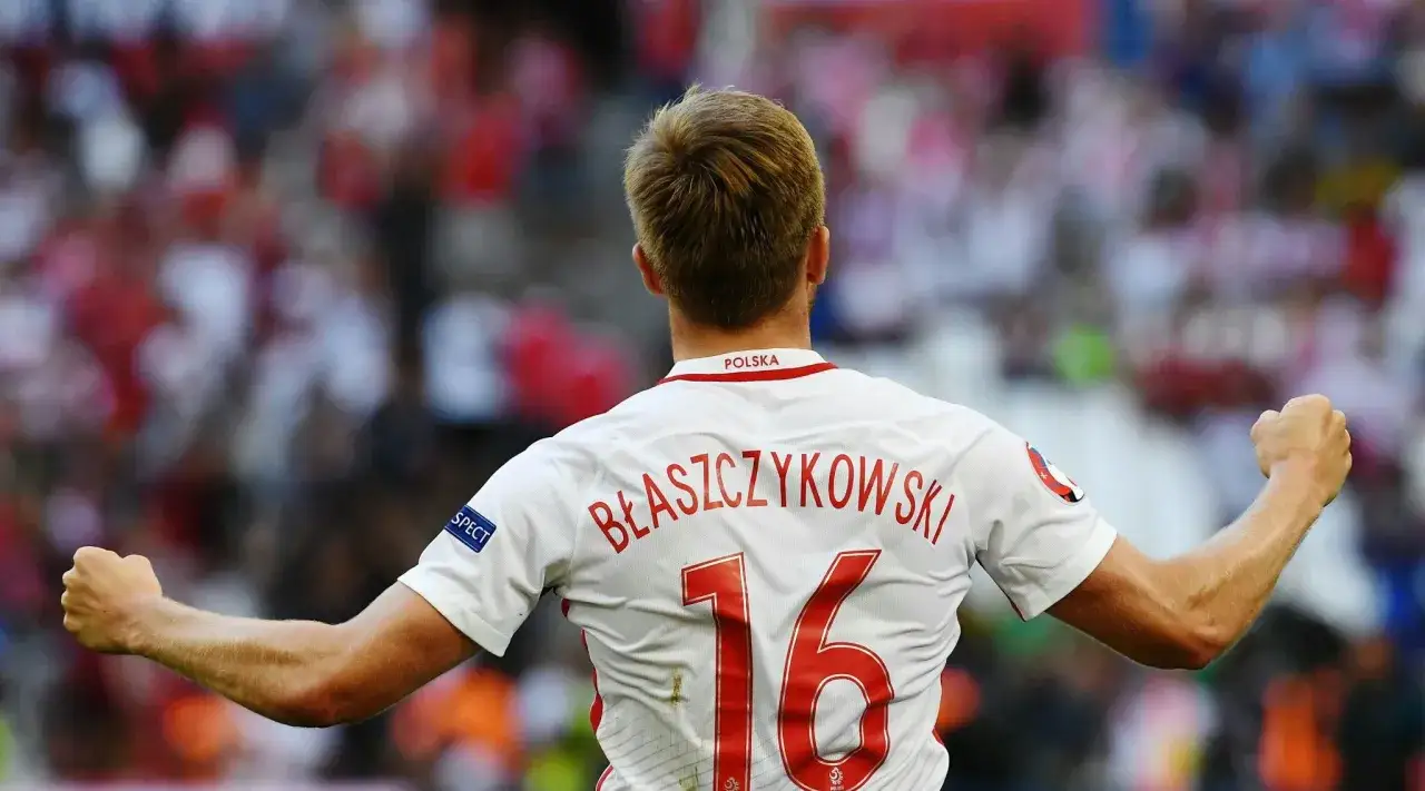 Piłkarz Jakub Błaszczykowski w koszulce z numerem 16, z podniesionymi pięściami. Jego majątek to wynik ciężkiej pracy na boisku.