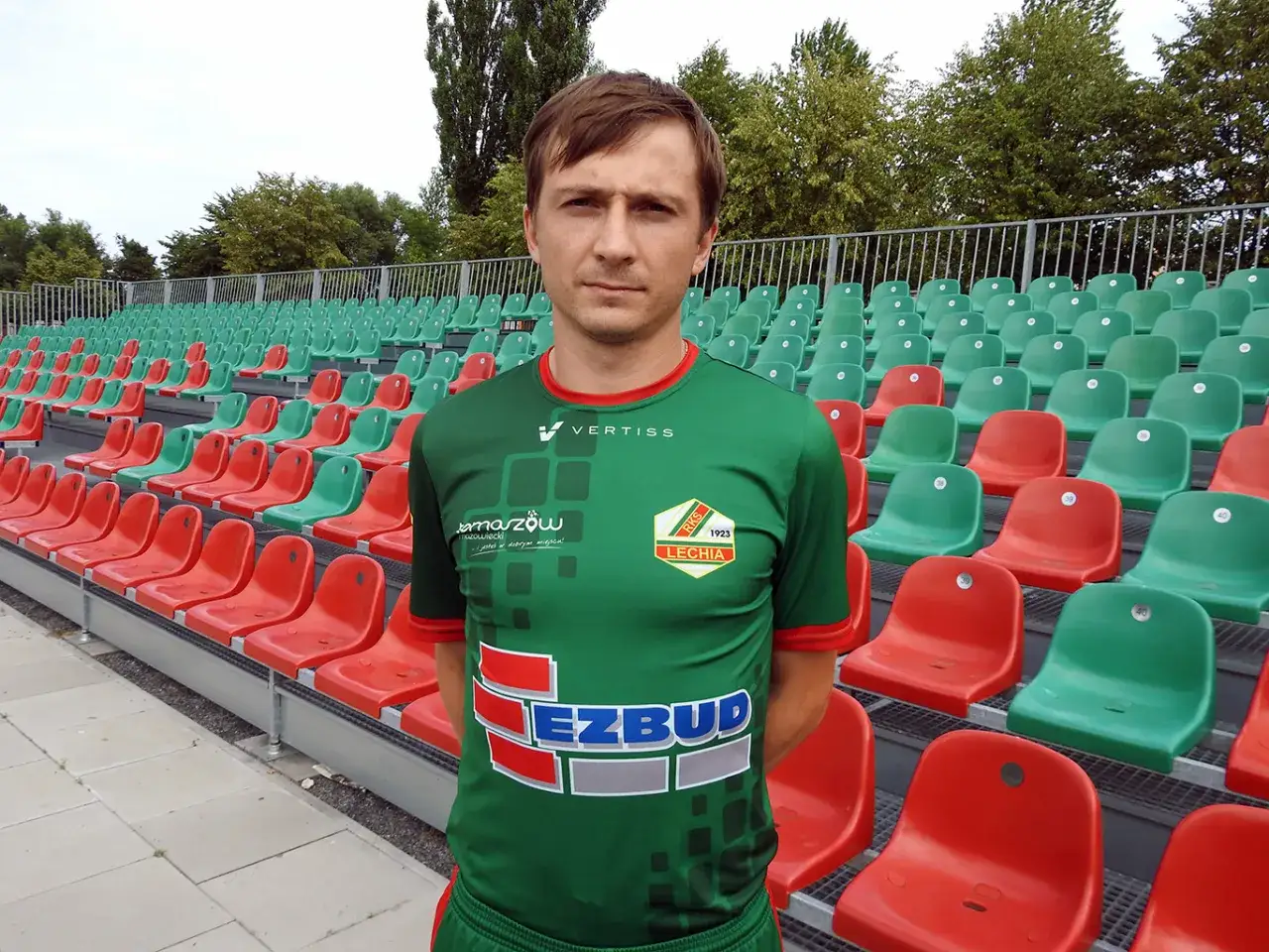 Kamil Szymczak w zielonej koszulce z herbem klubu na tle pustych, czerwono-zielonych trybun stadionu.