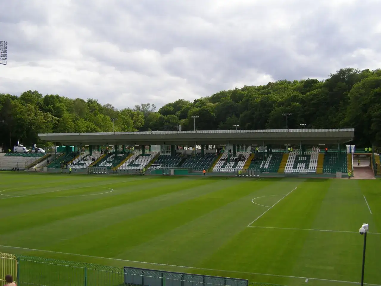 Stadion w Gdańsku, gdzie grają piłkarze. Zielona murawa, trybuny z białymi i zielonymi siedzeniami, las w tle.
