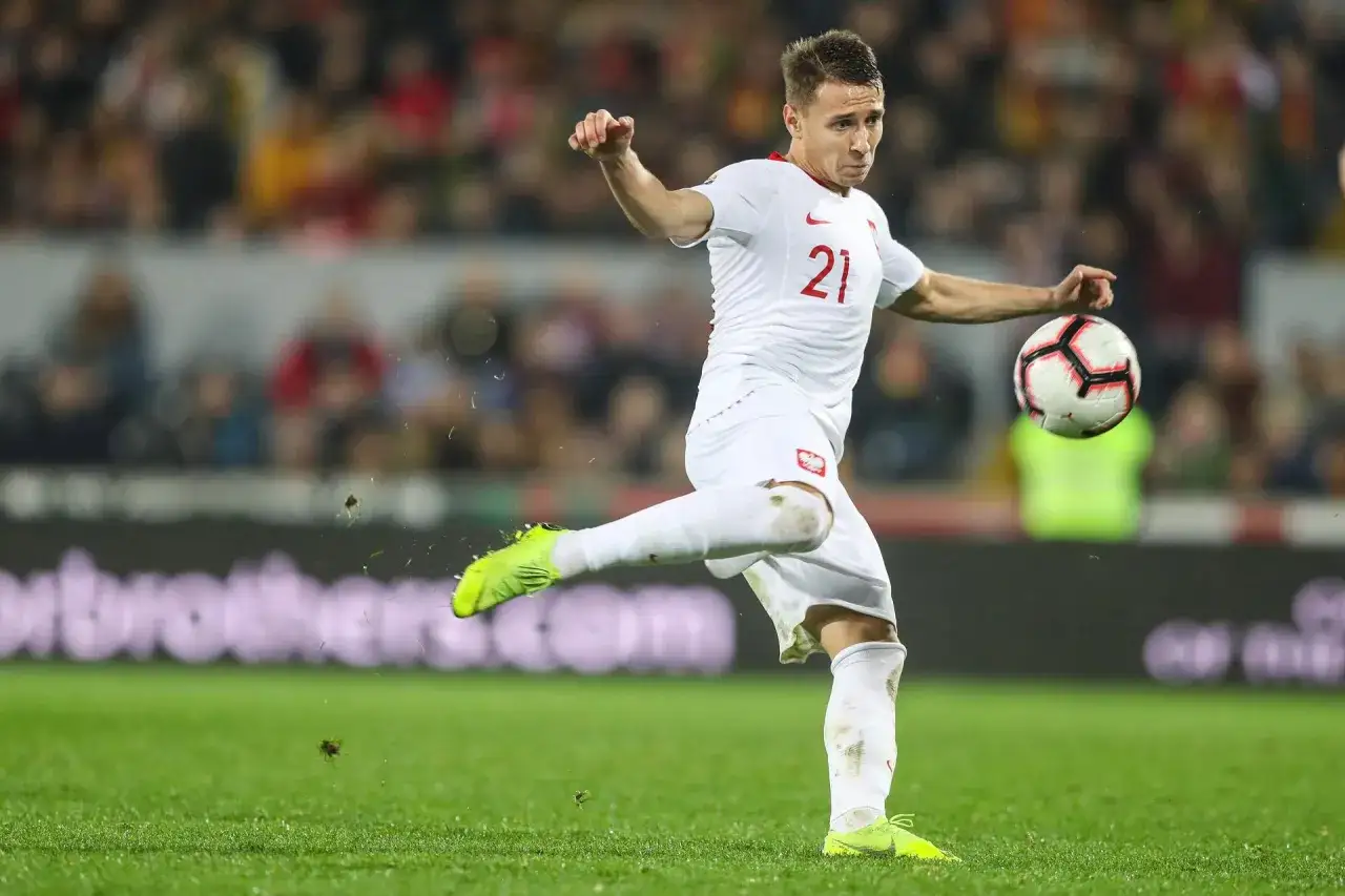 Frankowski piłkarz w akcji, w białej koszulce z numerem 21, kopie piłkę na zielonej murawie.