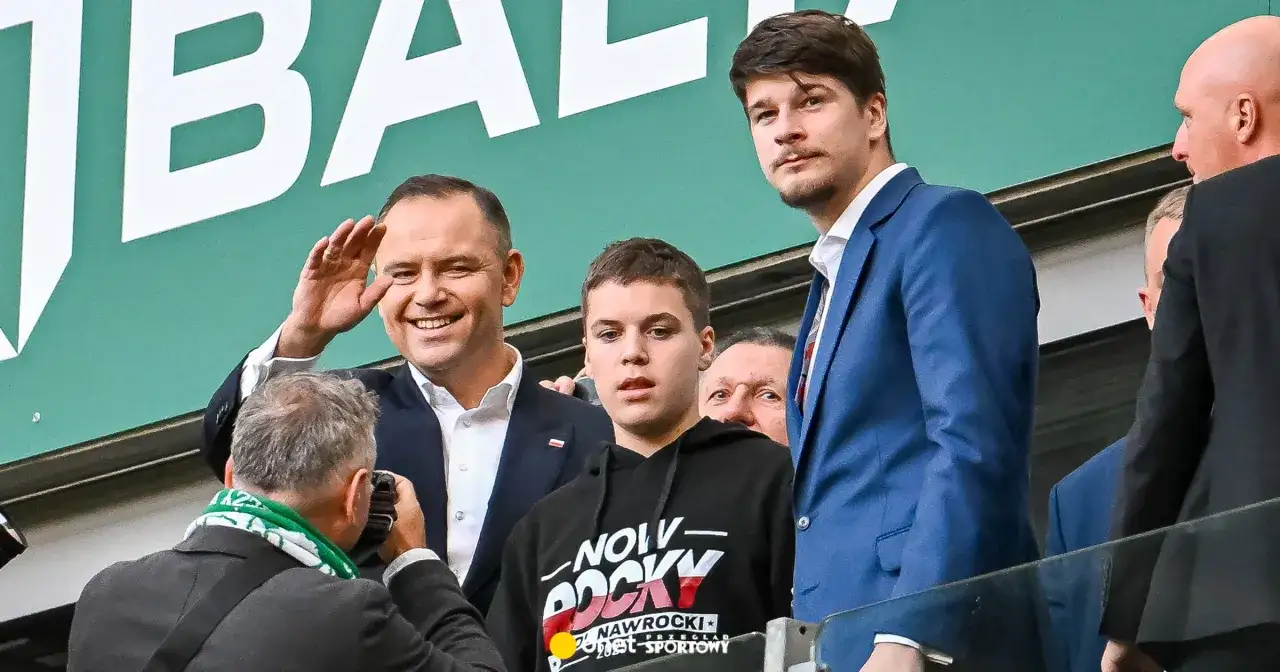 Mężczyzna w garniturze pozdrawia, obok młody chłopak w bluzie z napisem 