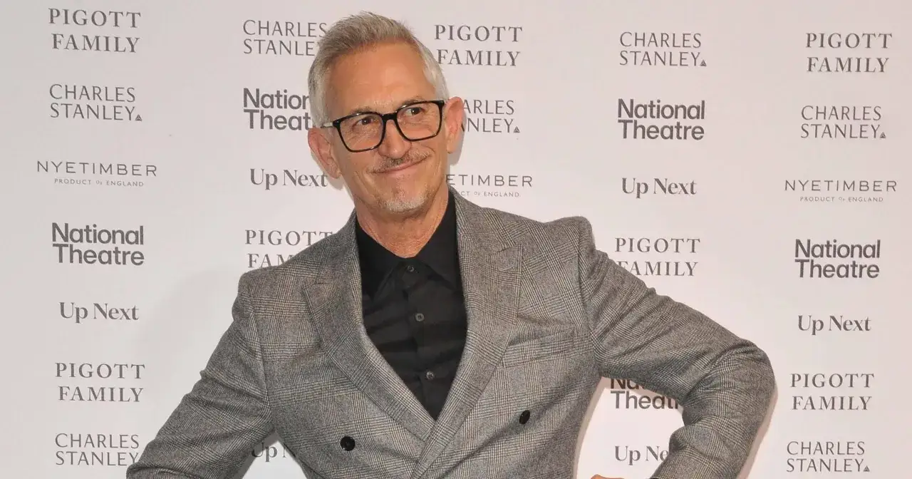 Gary Lineker w szarej marynarce z wzorem w kratę, z okularami na nosie, uśmiecha się na tle ścianki z logo National Theatre.