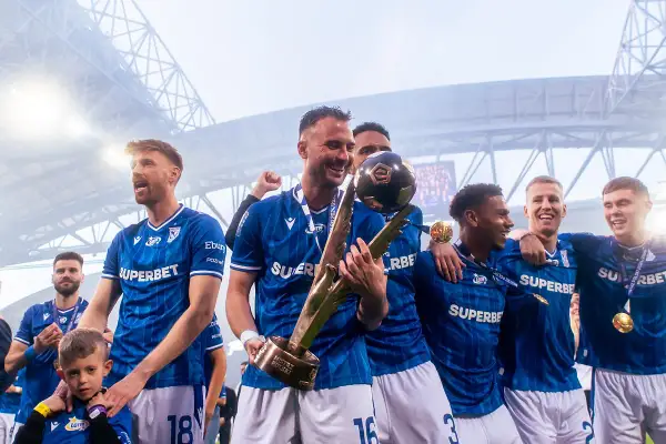 Piłkarze w niebieskich strojach świętują z pucharem i piłką na stadionie.
